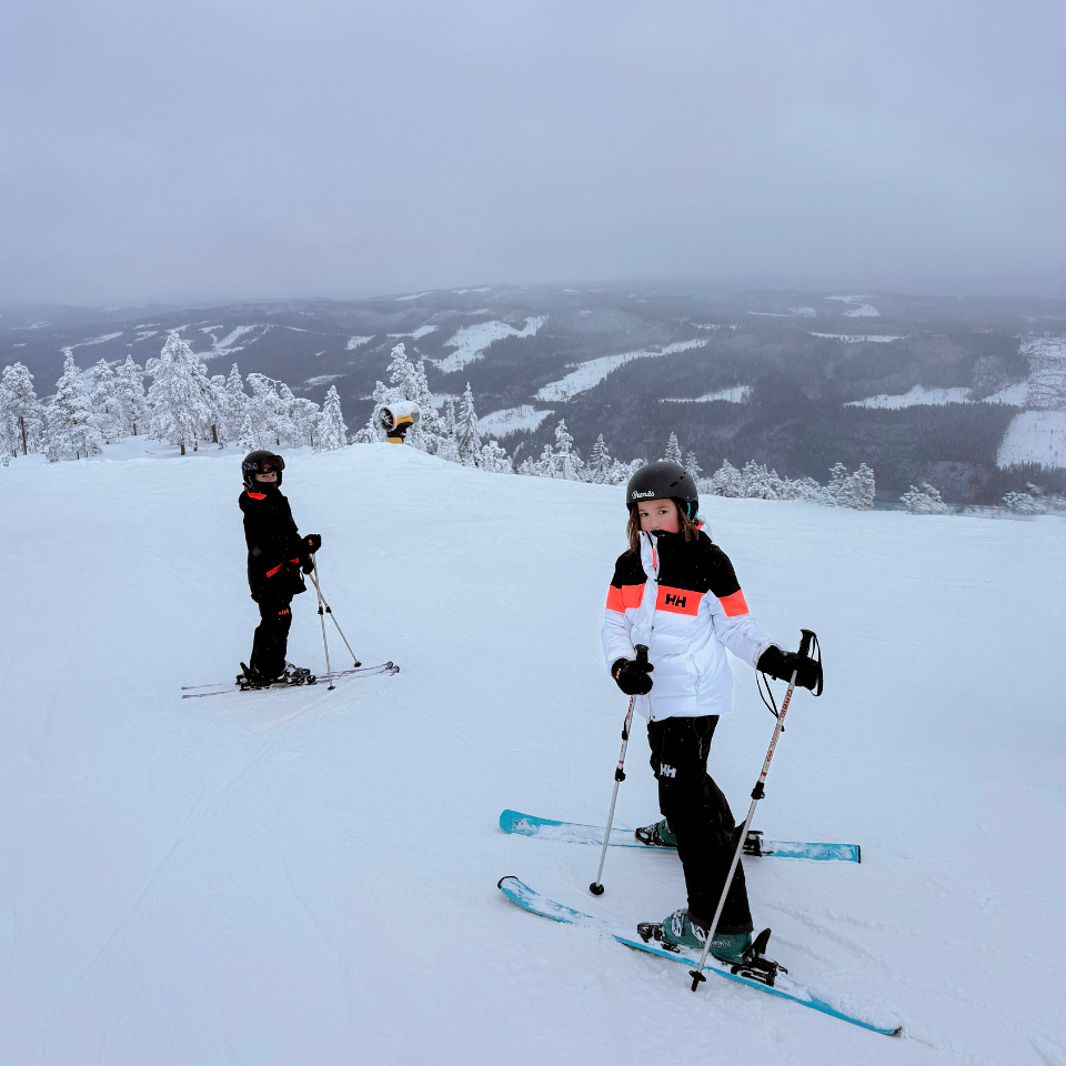 To børn på ski på en bred, velpræpareret piste i Branäs med sneklædt natur i baggrunden.