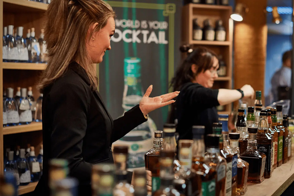 En kvindelig bartender præsenterer et udvalg af flasker ved en cocktailbar med teksten "The world is your cocktail".